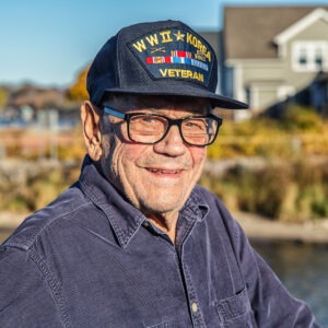 An elderly man with a WW 2 Veterans hat on smiles in front of a coastal New England scene.