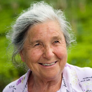 An elderly woman with gray hair smiles against a green, lush background